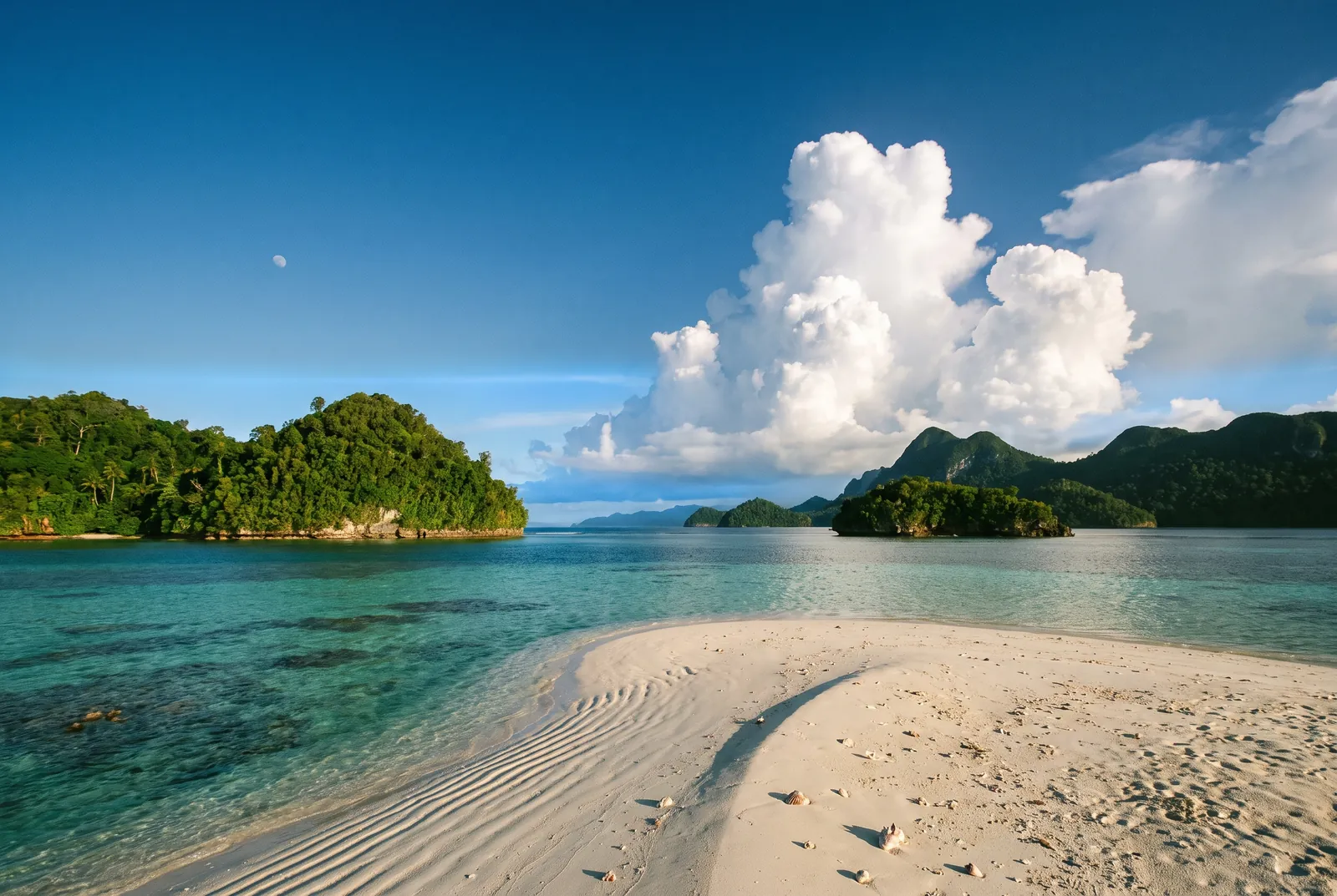 Barre de sable blanc au bord d’un lagon turquoise, îles couvertes de forêt et grands cumulus.